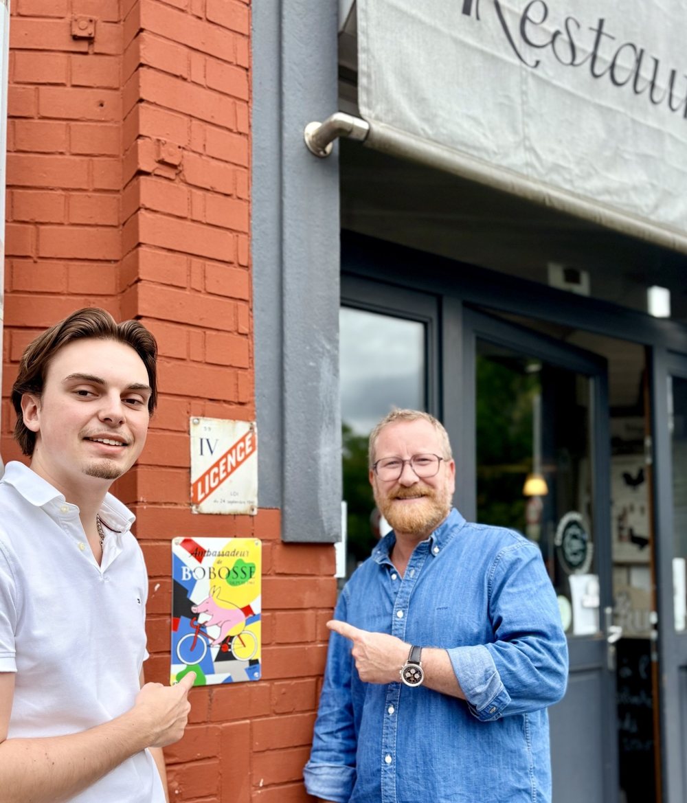 Jules et Alex montrent du doigt la plaque Ambassadeur Bobosse sur la façade du Triez, à côté de la plaque Licence IV