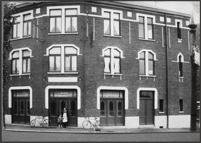 Photo ancienne en noir et blanc de la maison au coin du boulevard de la Marne à Mouvaux, avec l'enseigne J. Delbosse, deux personnes devant la porte et des vélos