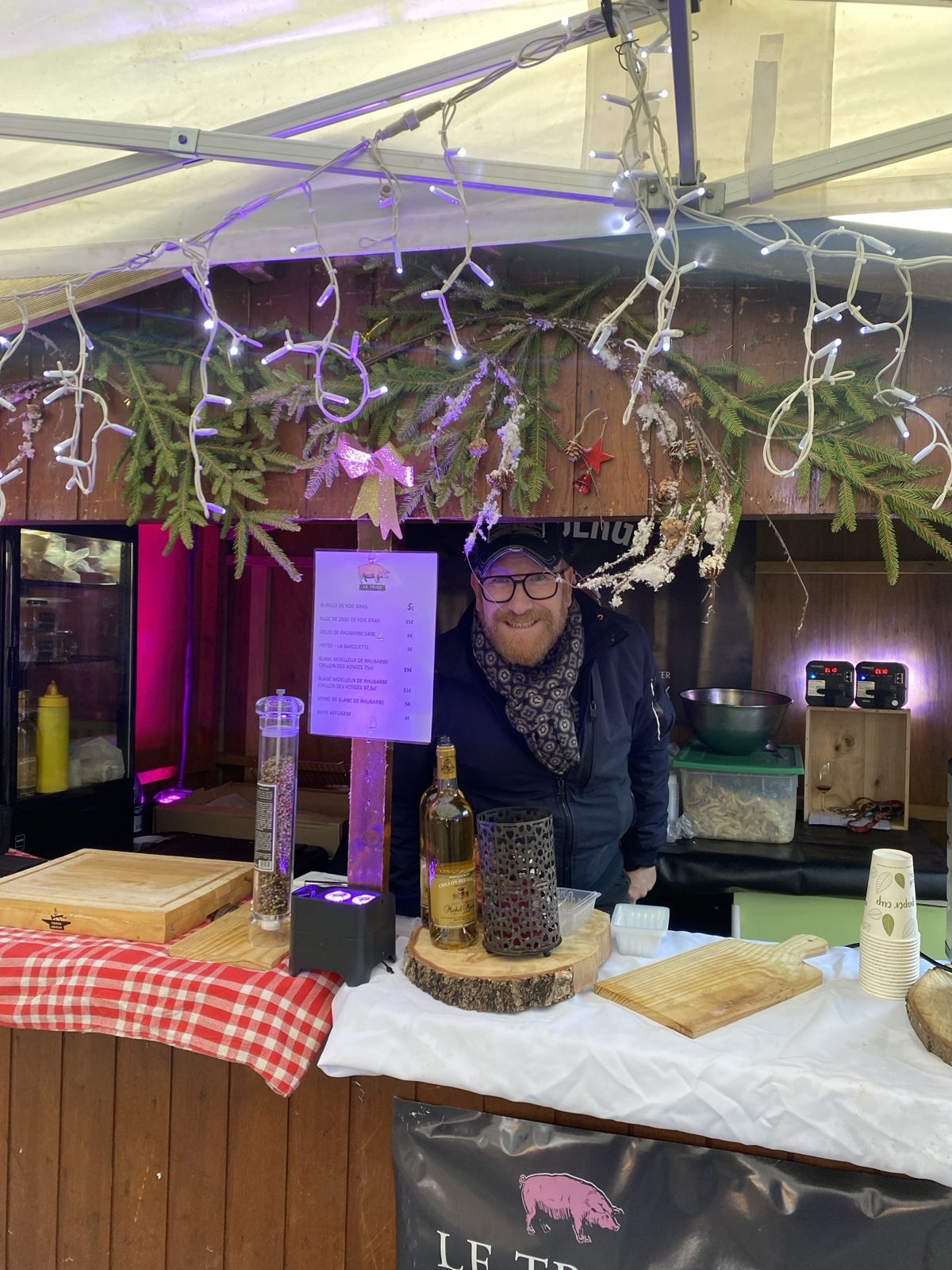 Alex souriant derrière le chalet du Triez, décoré de sapin et de guirlandes lumineuses, au marché de Noël de Mouvaux